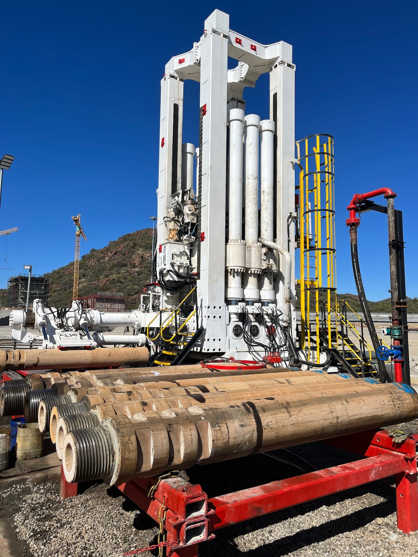 An industrial drilling rig stands tall against a clear blue sky with a hillside visible in the background. Thick cylindrical pipes are laid out on a red metal structure in the foreground. The rig has multiple large pistons and an array of interconnected machinery.