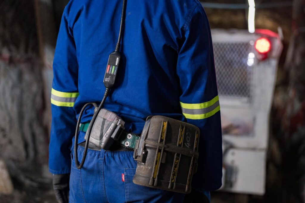 A worker in blue protective clothing with reflective yellow stripes and equipment attached to their belt is shown from behind. The worker is inside what appears to be a cave or underground tunnel, with machinery and dim lighting in the background.