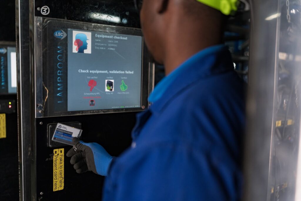 Person wearing blue protective clothing and a neon yellow hard hat checking equipment status on a screen using a card. The screen displays graphics and text for equipment checkout with indicators highlighting 'validation failed' in red.