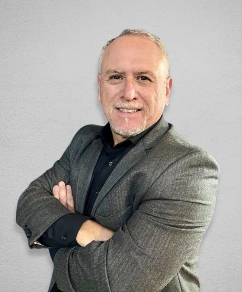 A man with short gray hair and a goatee is smiling at the camera. He is wearing a gray blazer over a black shirt, and his arms are crossed. The background is a simple light gray wall.