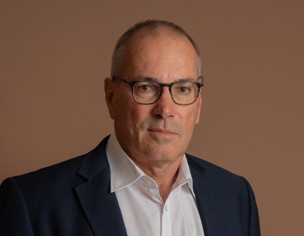A middle-aged man wearing glasses, a white shirt, and a dark blazer poses against a plain, light brown background. He has short hair and a neutral expression.