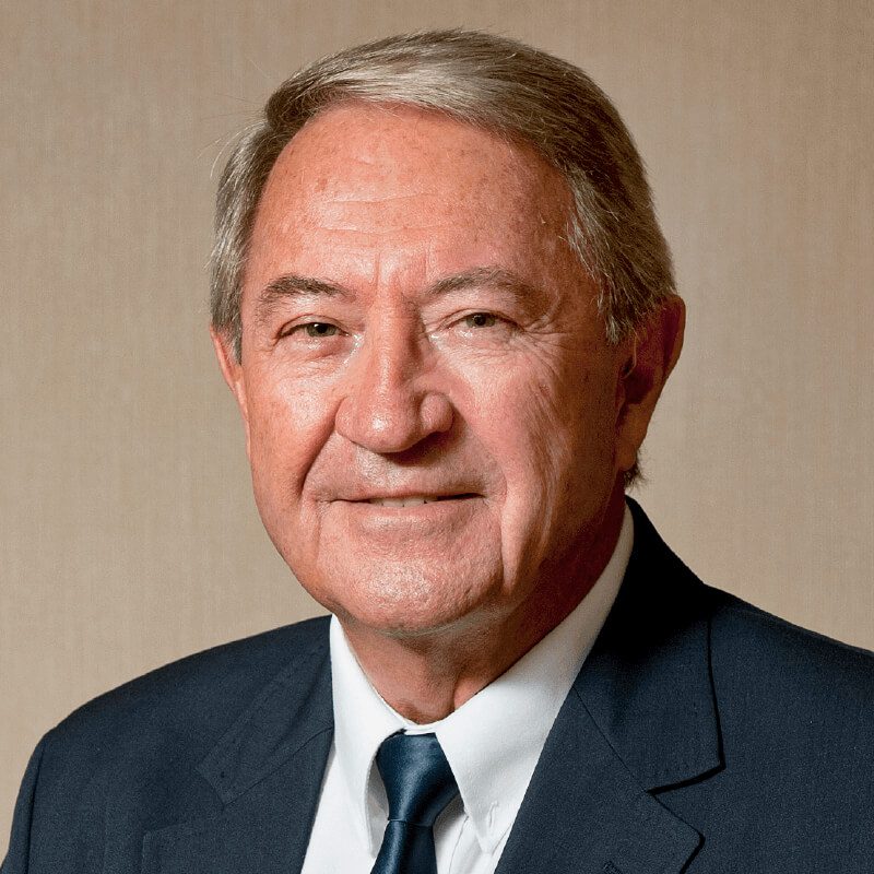 A senior man with short gray hair smiles slightly while looking at the camera. He is wearing a dark suit jacket, a white shirt, and a dark tie. The background is a plain beige wall.