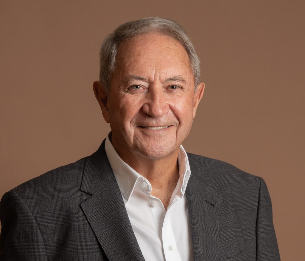 An older man with short gray hair is smiling at the camera. He is wearing a white dress shirt and a dark gray suit jacket, posed in front of a plain light brown background.