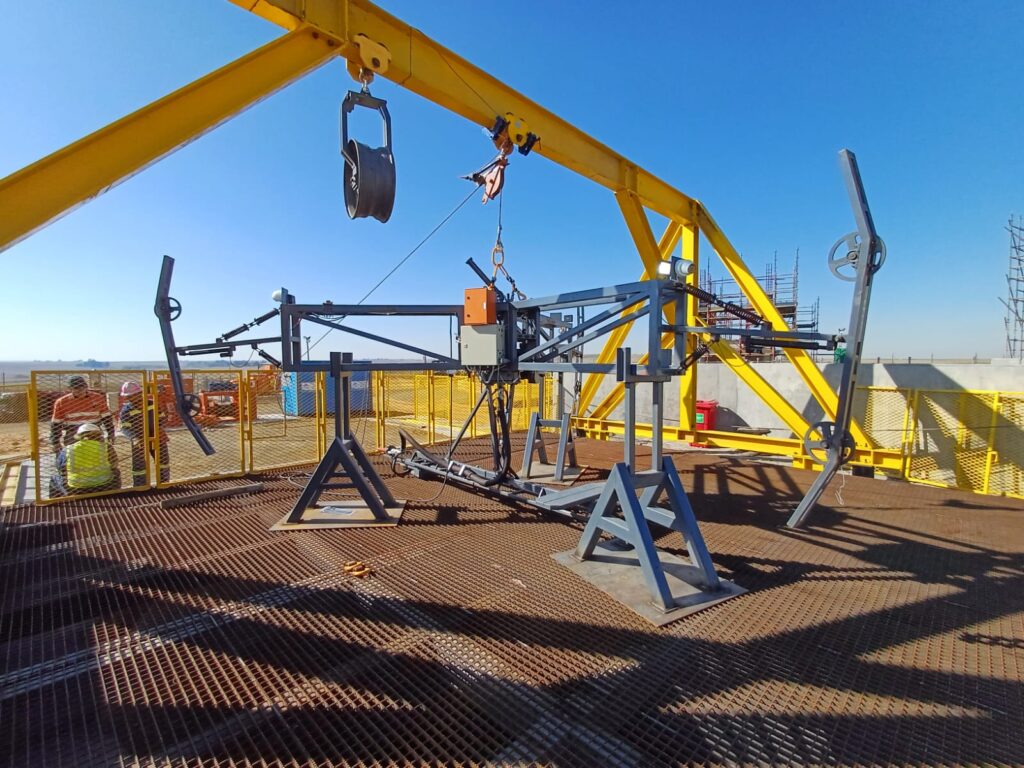 A metal testing rig on a grated platform under a bright blue sky. The setup includes a yellow gantry crane and multiple metallic components and wires. Construction materials and workers are visible in the background.