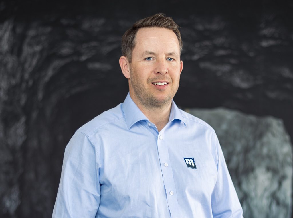 A man with short brown hair wearing a light blue button-up shirt with a logo stands in front of a dark, textured background, smiling at the camera.