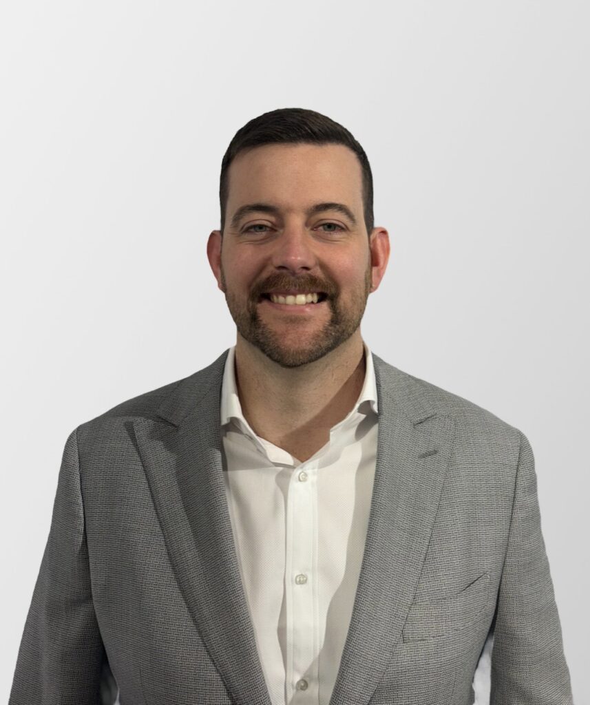 A man with a trimmed beard and moustache is smiling at the camera. He is wearing a light grey suit jacket over a white dress shirt. The background is plain and light grey.