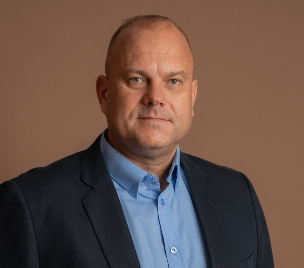 A man with short hair wearing a blue button-up shirt and a dark blazer poses against a plain brown background, looking directly at the camera with a neutral expression.