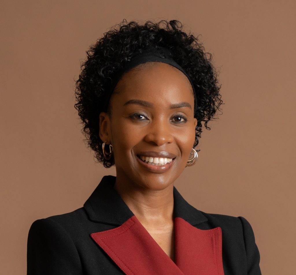 A woman with short curly hair smiles confidently. She is wearing hoop earrings and a black blazer with a maroon lapel, standing against a plain brown background.