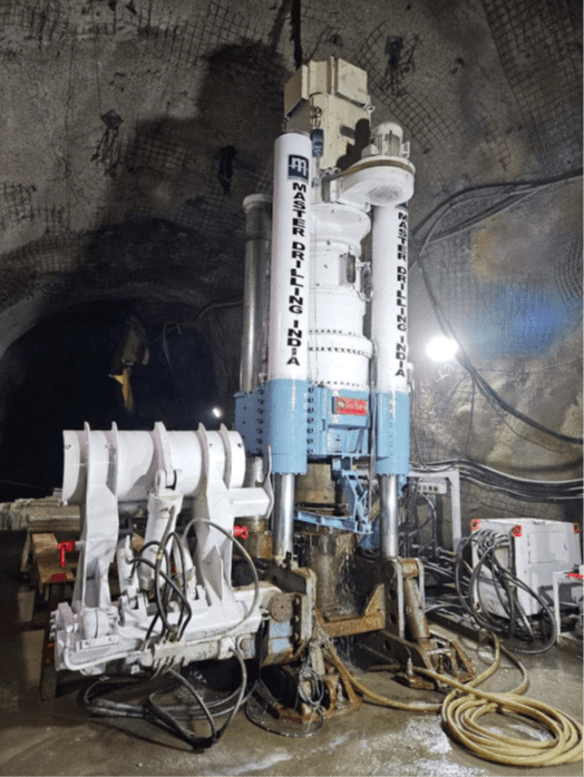 A large underground mining machine with two tall vertical components labeled "MASTER DRILLING." It is positioned in a dimly lit tunnel surrounded by cables and equipment on a wet concrete surface.