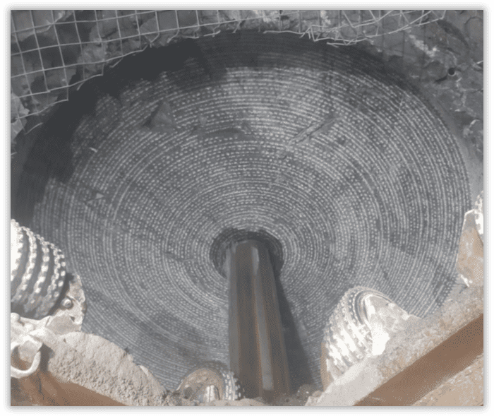 A circular tunnel boring machine viewed from the inside, showcasing concentric rings and the machine's drilling mechanism. The tunnel walls are grey, with a mesh visible at the top. Drilling heads frame the lower edges of the image.