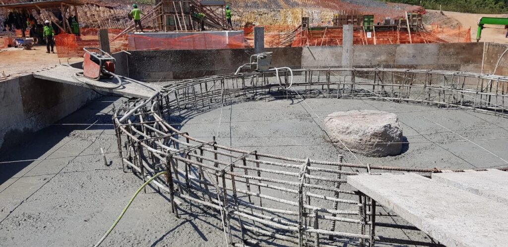 Construction site showing a circular concrete framework with steel rebar and partially poured concrete. Workers in safety gear stand nearby, with orange safety netting and equipment surrounding the area. A large rock is visible within the structure.