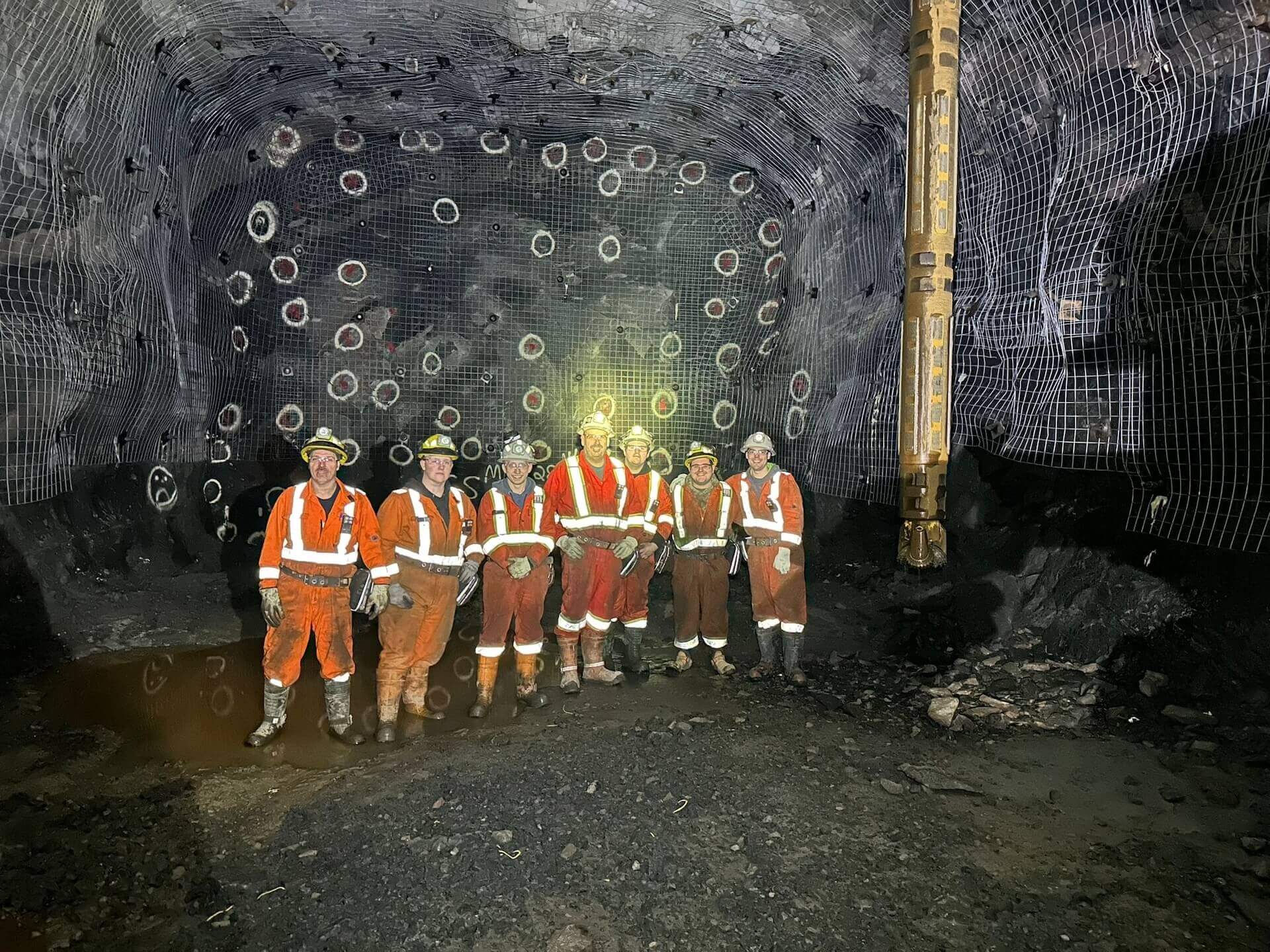 A group of construction workers wearing orange and red safety gear and helmets are standing together in an underground mine tunnel. The tunnel has a rocky, dark interior with illuminated grid patterns on the walls and various machinery in the background.