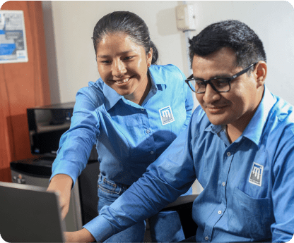 Two people, a woman and a man, dressed in matching company uniforms with a logo, are working together in an office. The woman is smiling and pointing at the computer monitor while the man is seated, looking at the screen with a focused expression.