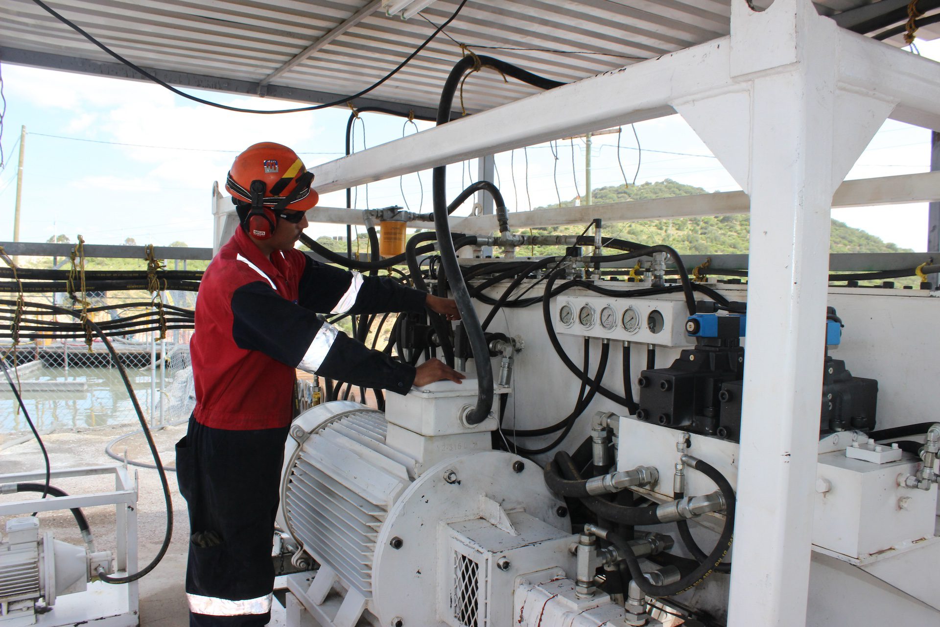 A worker wearing a red and black uniform, hard hat, and protective headphones operates machinery at an industrial site. The worker is adjusting a control on a large, white industrial motor, surrounded by various cables and equipment. Background shows an outdoor setting.