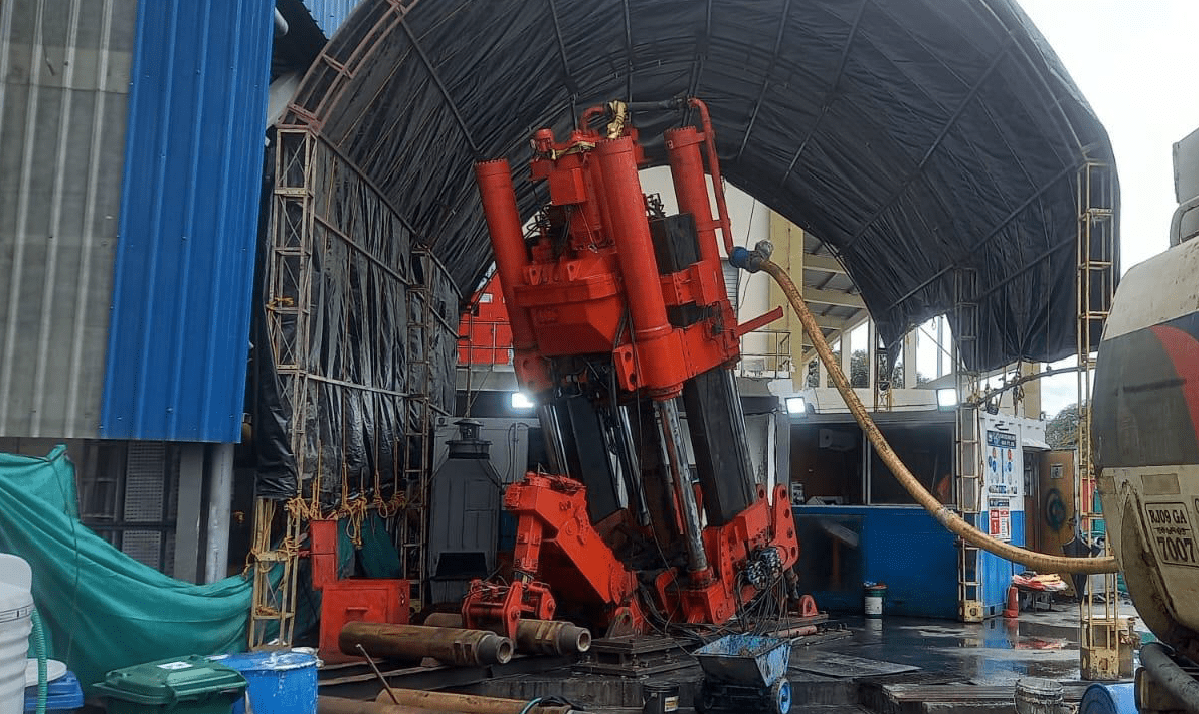 Industrial machinery under a large canopy, featuring tall red hydraulic components. The wet concrete floor and surrounding tools suggest maintenance or operation activity. The setting appears to be an outdoor industrial facility.