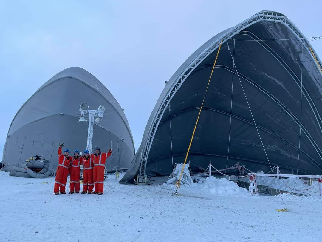 A group of five people wearing orange winter coats and hard hats stand on a snowy ground, raising their arms. They are in front of two large, semi-cylindrical, gray structures covered in snow. The sky is overcast.