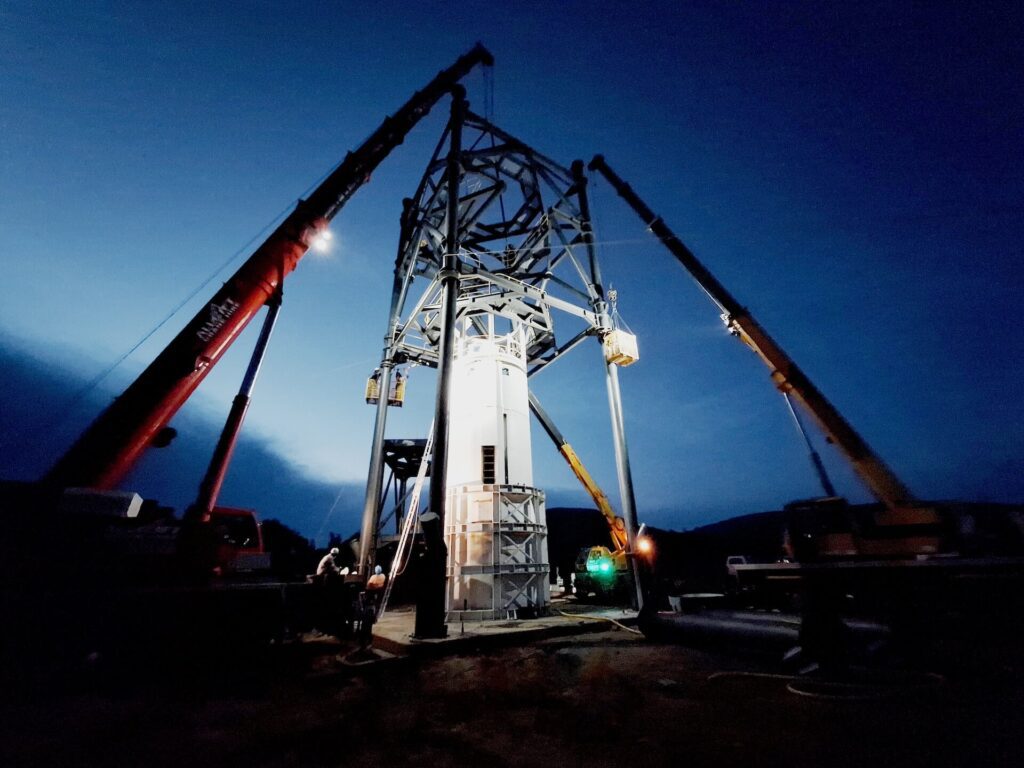 A large metal structure is illuminated against a twilight sky, with four cranes positioned around it, lifting and securing its components. Workers and equipment are visible at the base, indicating construction activity. The sky is a deep blue, nearing darkness.