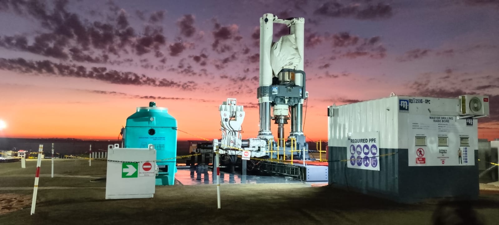 A construction site at sunset with a large piece of industrial machinery in the center. Nearby are a turquoise tank, a white container with safety signs, and a fenced-off perimeter. The sky is vividly colored with shades of orange, pink, and purple.