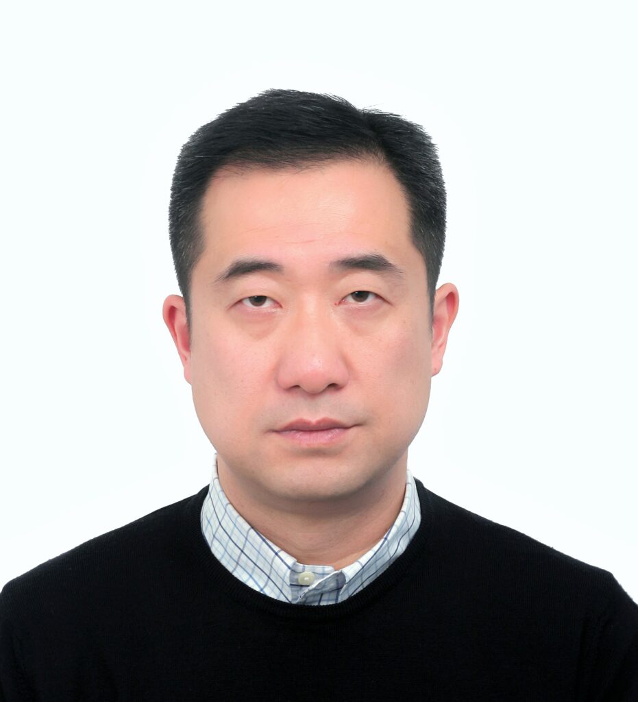 A man with short black hair is looking directly at the camera with a neutral expression. He is wearing a black sweater over a light-colored, checkered shirt. The background is plain white.