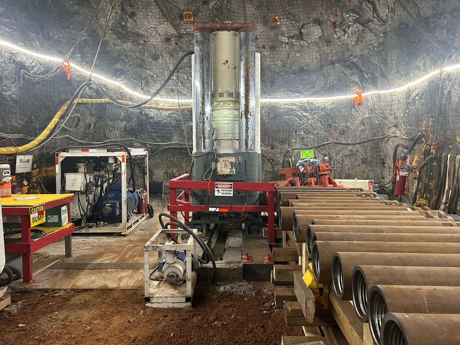 An industrial underground mining setup showing a drilling machine at the center, surrounded by various equipment and cables. To the right, several large cylinders are stacked. The background consists of rough rock walls. The area is lit with overhead strip lighting.