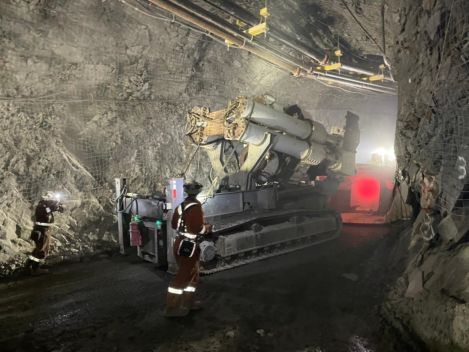 A large drilling machine operates inside a dimly lit underground mine tunnel, with two miners in safety gear standing nearby. One is holding a flashlight and inspecting the tunnel wall, while the other observes the machinery.