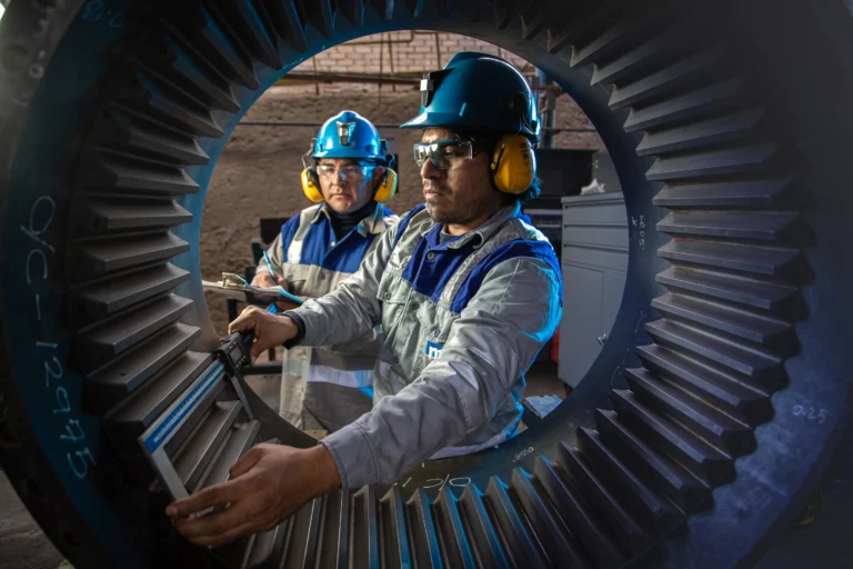 Two industrial workers with safety gear inspect a large gear mechanism. One worker, framed by the circular opening of the gear, measures its dimensions with a caliper while the other looks on. Both wear helmets, safety glasses, and ear protection in a workshop setting.