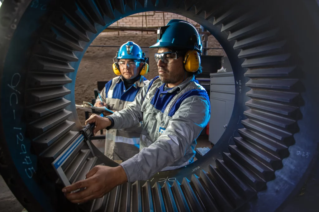 Two workers in safety gear, including helmets, ear protection, and safety glasses, inspect a large, industrial gear. One worker uses a measuring tool, while the other observes, both framed by the gear's teeth, emphasizing precision and teamwork in an industrial setting.