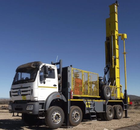 A large drilling rig mounted on a heavy-duty truck, parked on a barren, rocky terrain. The truck is white and steel-gray, while the vertical drilling apparatus is bright yellow. The clear blue sky contrasts with the rugged landscape in the background.