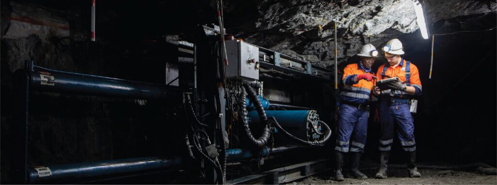 Two miners wearing helmets and safety gear stand in a dark underground tunnel illuminated by a single light. They are working with machinery and appear to be coordinating or checking something on a tablet. The background is rugged with rock and mining equipment.