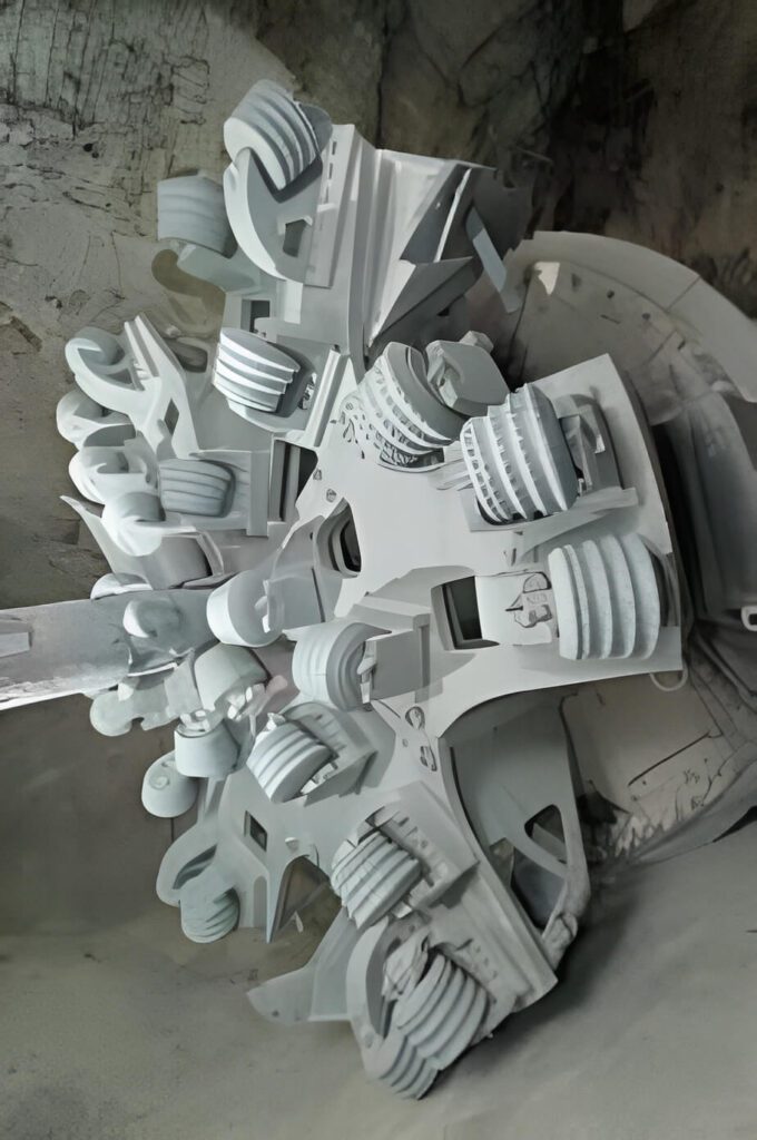 A modern tunnel boring machine with intricate mechanical parts and rotating cutters is positioned underground. The grey and metallic apparatus is designed for excavation, with several large circular cutting heads and heavy-duty components visible.