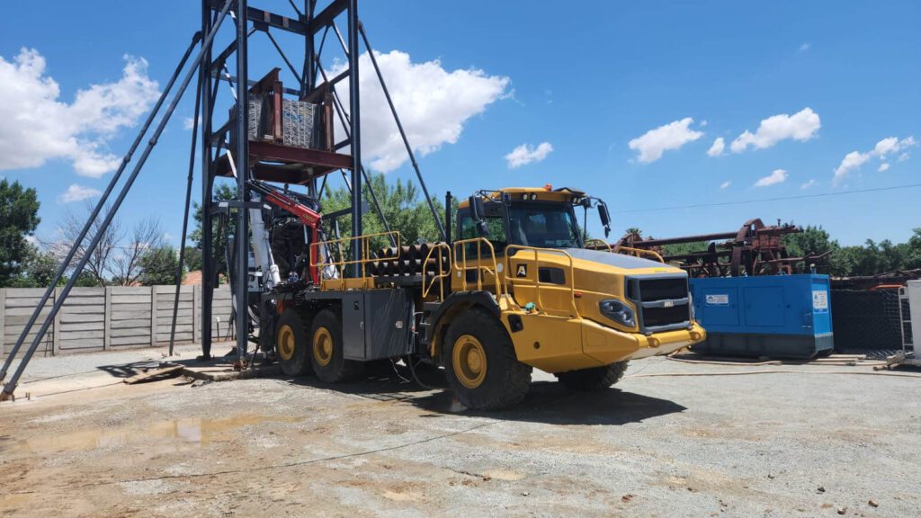 A large yellow construction vehicle is positioned near a tall, metal framework structure in an outdoor work area. The clear blue sky is dotted with a few clouds, and various equipment and materials are scattered around the site.