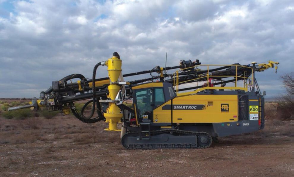 A large, yellow drilling machine labeled "SMART ROC" is positioned on rough terrain under a cloudy sky. The vehicle features multiple hydraulic arms and equipment for mining or construction use. Number "600" is displayed on a tag near the front.