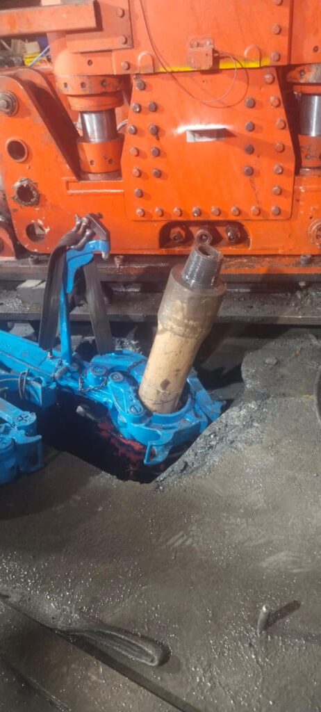 A mechanical arm grips a cylindrical object, in front of an orange industrial machine. The area around the machine appears to be a dusty or dirty workshop floor. The scene likely depicts part of a manufacturing or repair process.