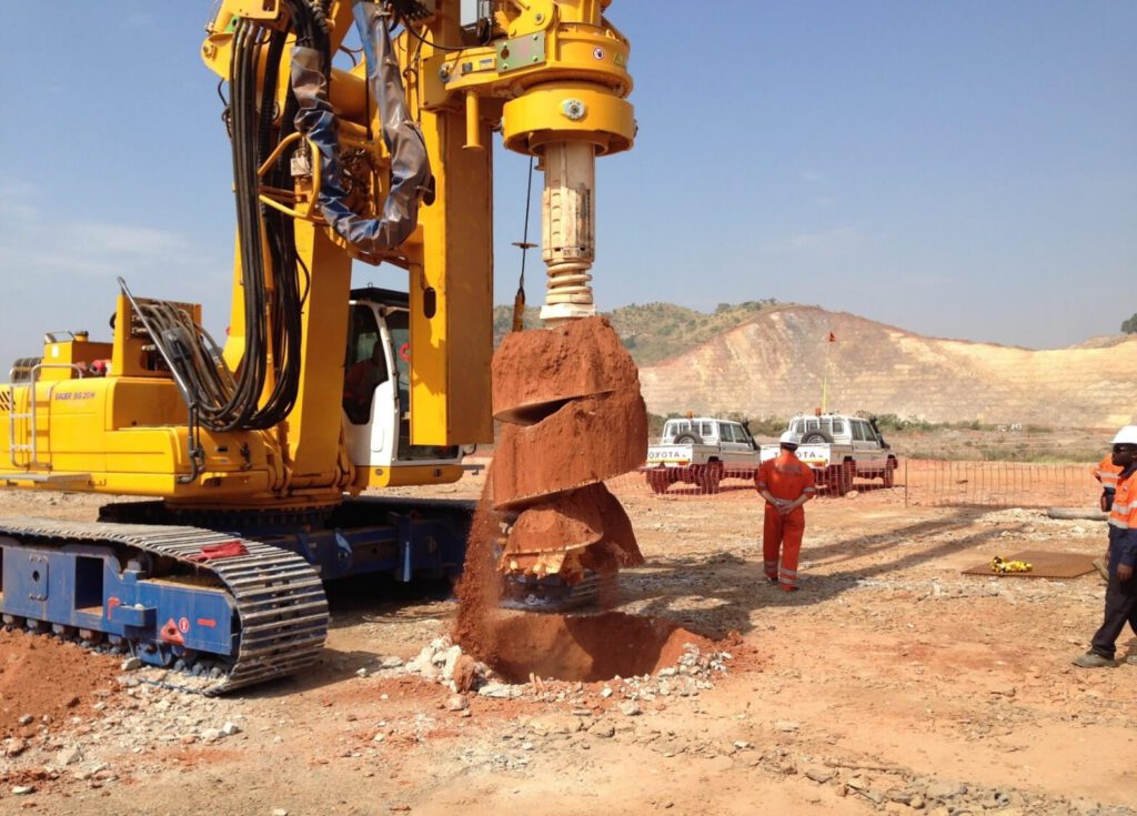 A large yellow drilling machine with a spiral auger attachment is in operation at a construction site. Workers in orange safety gear stand nearby, overseeing the work. The background features a hilly landscape and several utility vehicles.