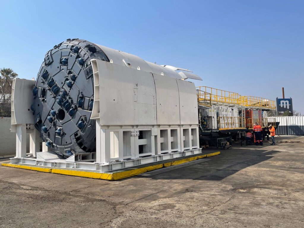 A massive tunnel boring machine is stationed on a flat surface outdoors. The front cutting head is round with multiple cutting blades and is positioned horizontally. Several workers in high-visibility clothing stand nearby next to a control unit and various machinery.