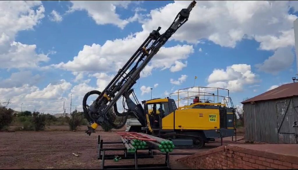 A bright yellow drilling machine with an extended drill is stationed on a dry, dirt ground under a partly cloudy sky. Nearby, green cylindrical objects are placed on a metal rack, and a wooden structure is visible to the right.