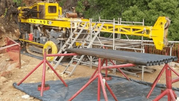 A yellow hydraulic drilling rig is set up on a construction site, surrounded by scaffolding and safety barriers. The ground is covered with tarpaulin and metal pipes are arranged on a red support frame in front of the machine. Green trees are visible in the background.