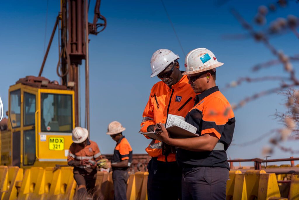 Workers in safety gear, including hard hats and high-visibility clothing, are engaged in outdoor activities at a construction site. Two are reviewing documents, while another operates heavy equipment in the background under a clear blue sky.