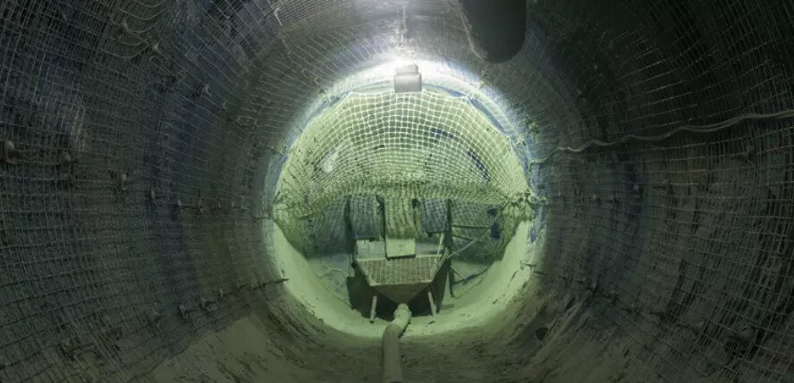 A dimly-lit underground tunnel reinforced with metal netting on the walls and ceiling. The floor is rough and appears to have construction equipment and debris scattered around, suggesting an ongoing or recently completed excavation project.