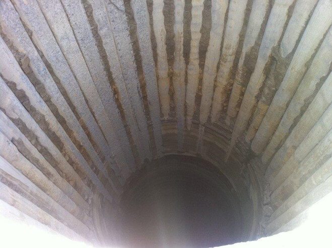 A deep, circular well with ribbed, stone walls extending down into darkness. The light at the opening casts shadows, emphasizing the textured surface and the well's depth.