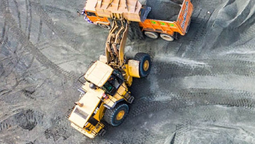 An aerial view of a construction site shows a yellow excavator loading dirt into an orange dump truck. The vehicles are on a rough, gray, dirt surface with tire tracks visible around them. The excavator's arm is extended into the back of the dump truck.