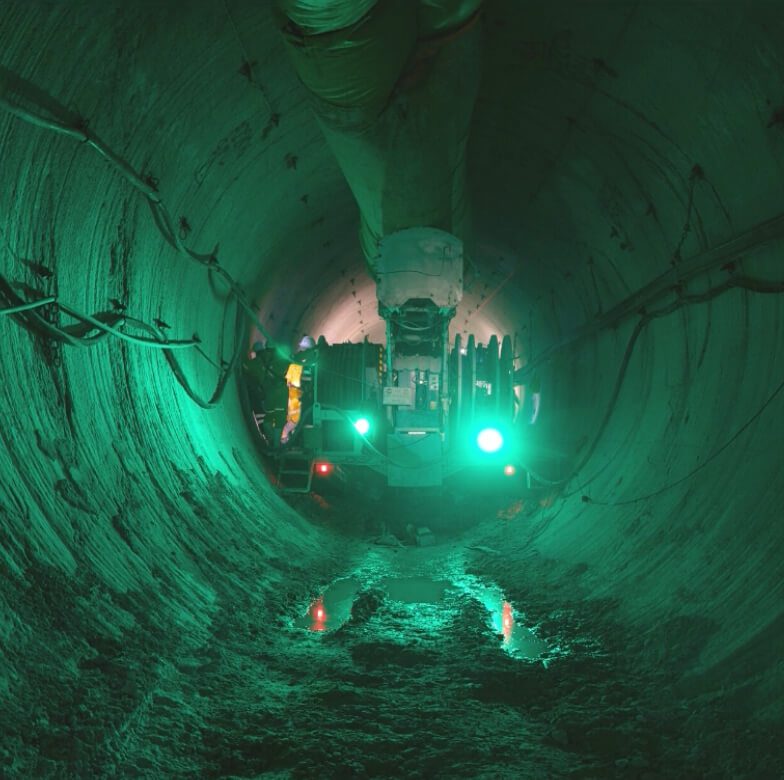 A worker operates machinery inside a large, illuminated tunnel with rugged, uneven walls. Green lighting casts an eerie glow, and there is a small puddle of water on the ground reflecting the lights. The scene appears industrial and underground.