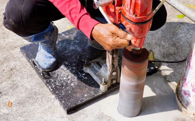 A worker operates a core drill to bore a hole into a concrete surface. One hand grips the handle while the other steadies the equipment. The worker wears a red long-sleeve shirt and blue protective boots. The equipment is mounted on a black base.