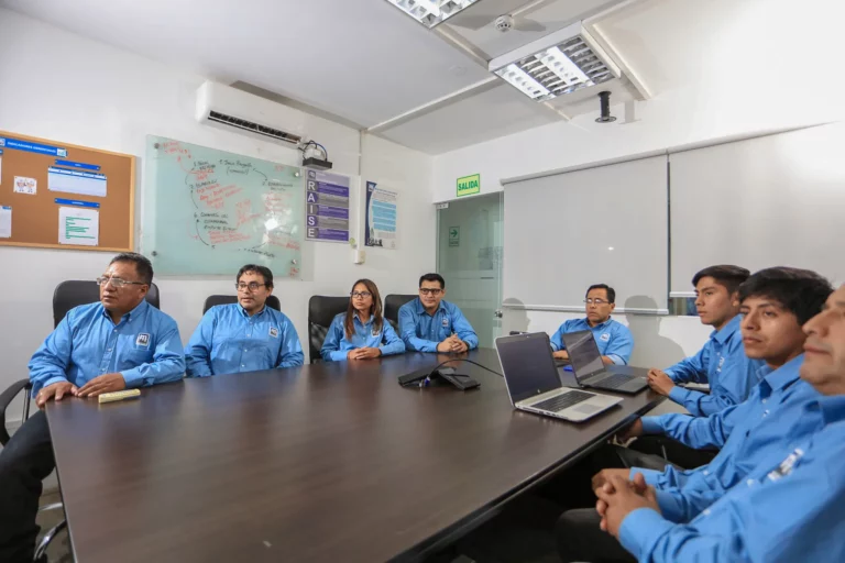 A group of eight people in blue uniforms sits around a long conference table in a well-lit room. The table has laptops and documents. A whiteboard and a bulletin board with notes are visible in the background. The people appear to be engaged in a meeting.