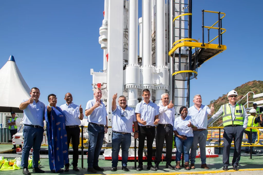 A group of nine people, some in professional attire and others in safety gear, stand in front of a large rocket-like structure. They are all smiling and giving a thumbs-up, with a clear blue sky and a hill in the background.