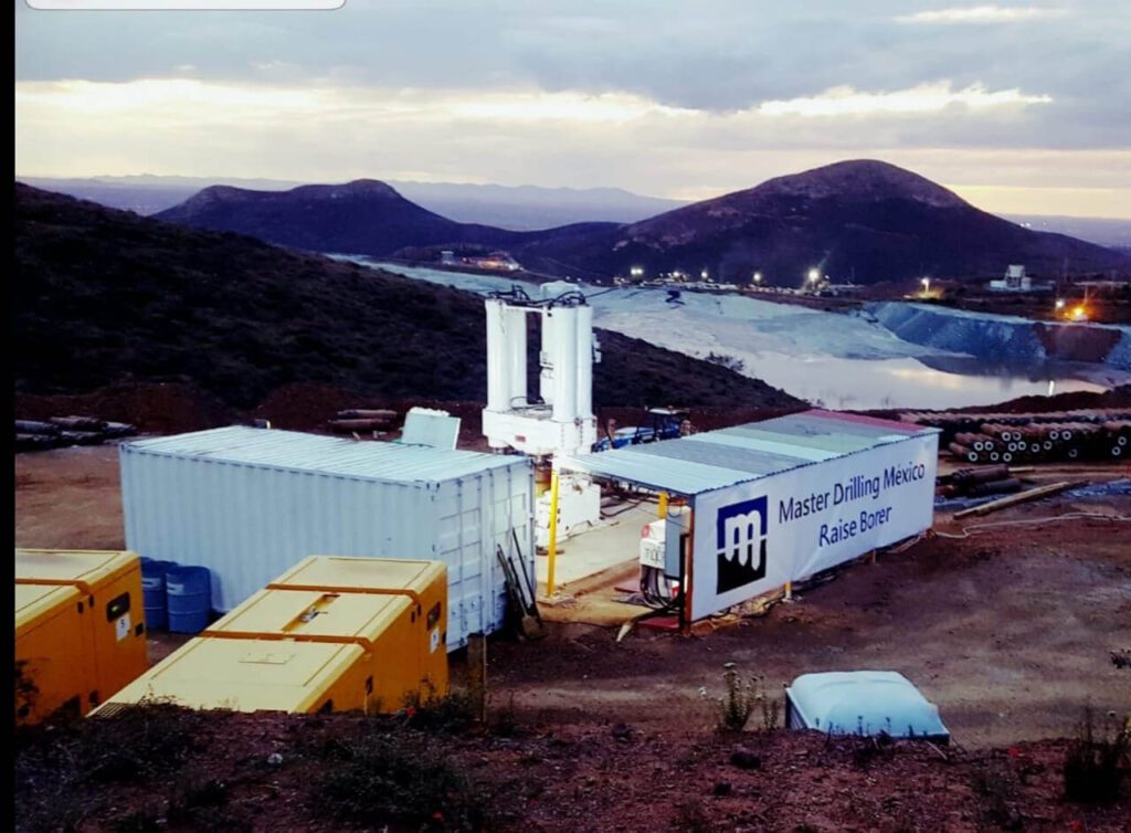 An industrial setup in a mountainous area at dusk, featuring a white tower-like structure, container units, and yellow machinery. A sign on one container reads "Master Drilling Mexico Raise Boring." In the background, hills and a large, lighted pit area are visible.