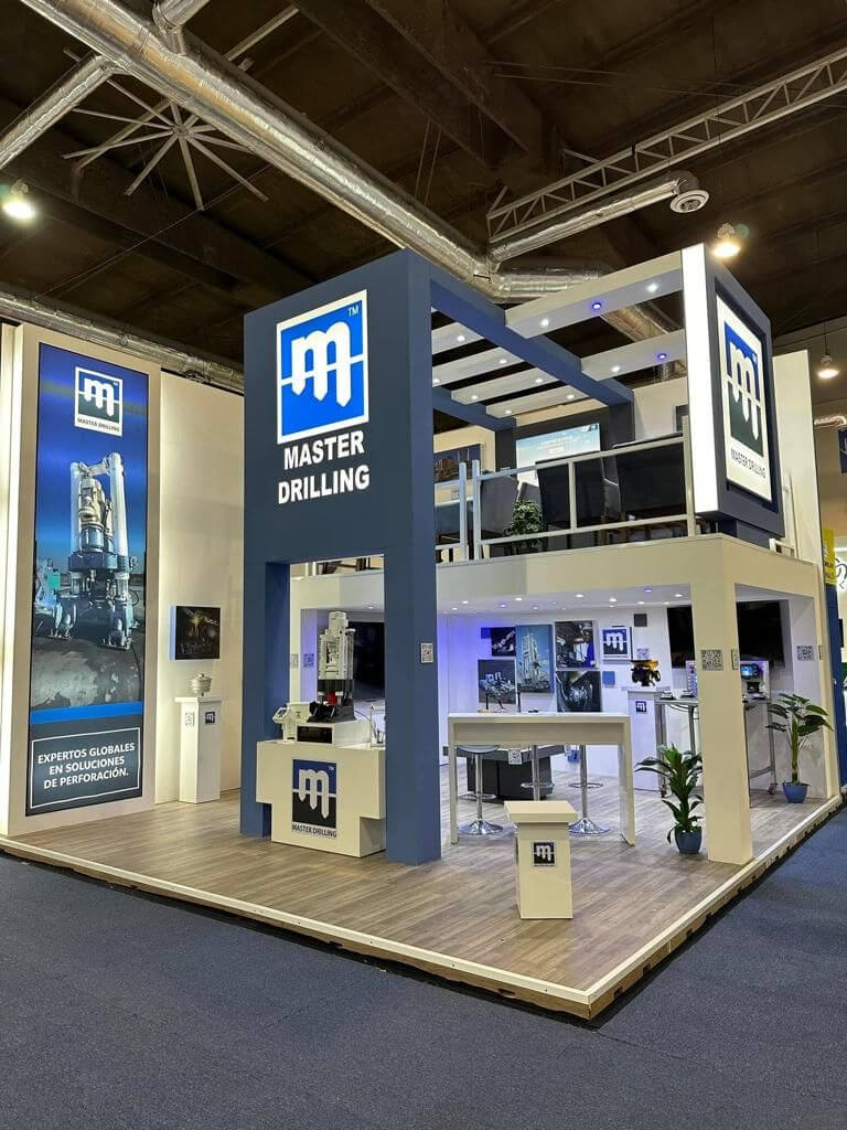 A two-story exhibition booth for Master Drilling, featuring a modern design with blue and white colors, illuminated signage, and promotional displays. There's a seating area, informational posters, and a small plant. The logo and branding are prominently displayed.