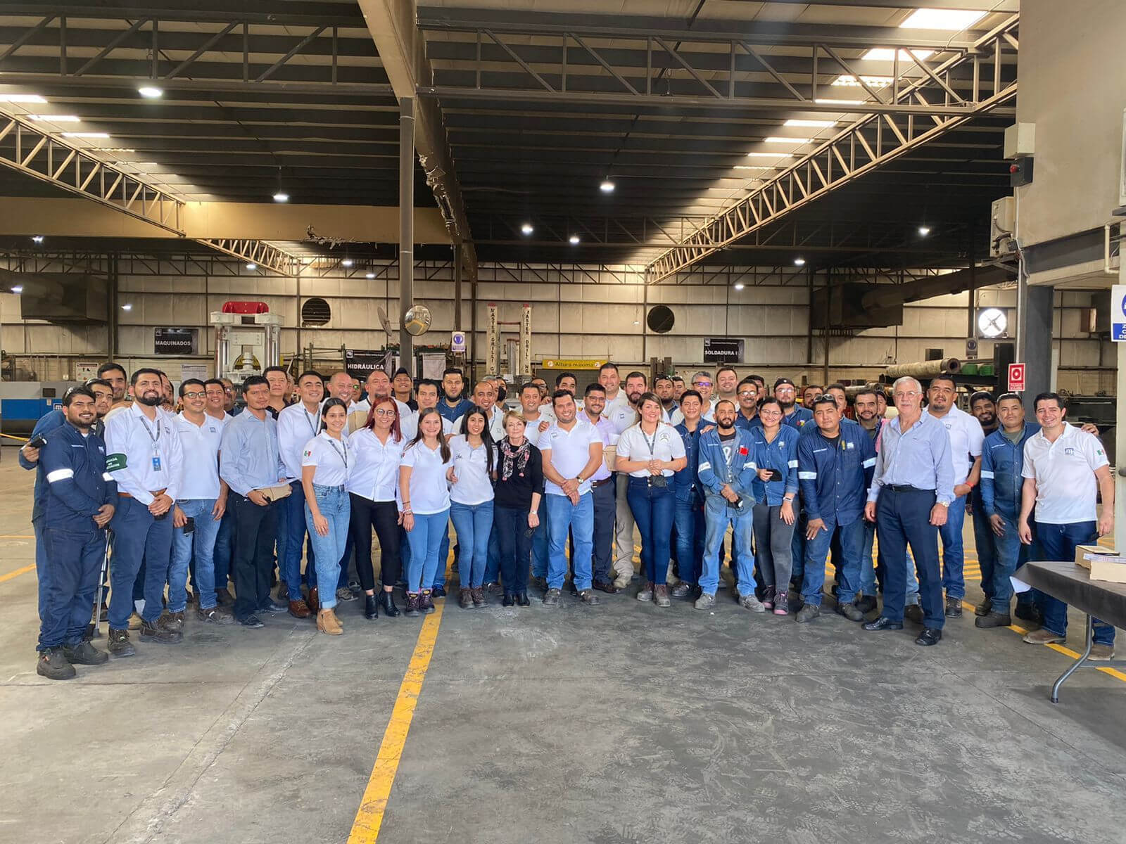 A large group of people poses for a photo inside an industrial warehouse. The group consists of men and women wearing casual business or work attire, including polo shirts, jeans, and uniforms. The warehouse features high ceilings, metal beams, and equipment shelves.