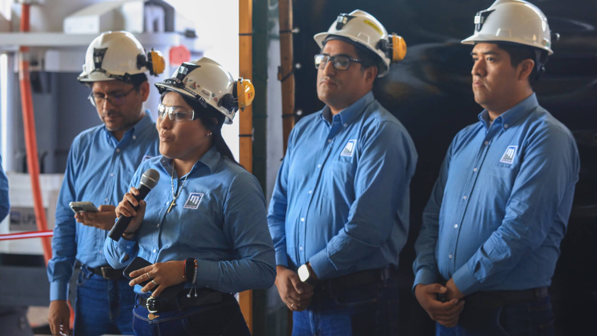 Four workers wearing blue shirts and white hard hats stand together. One worker, a woman, is speaking into a microphone while the others listen attentively. They appear to be in an industrial or construction setting.