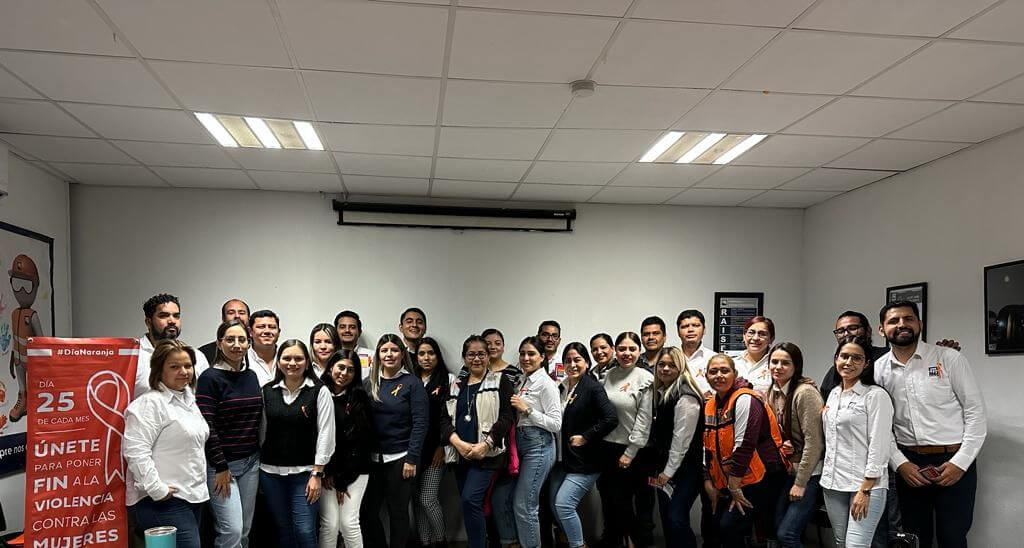 A group of people is standing together in a brightly lit room, smiling for a photo. To the left, there is a red banner with white text related to violence against women in Spanish. Everyone appears to be dressed in casual or business-casual attire.